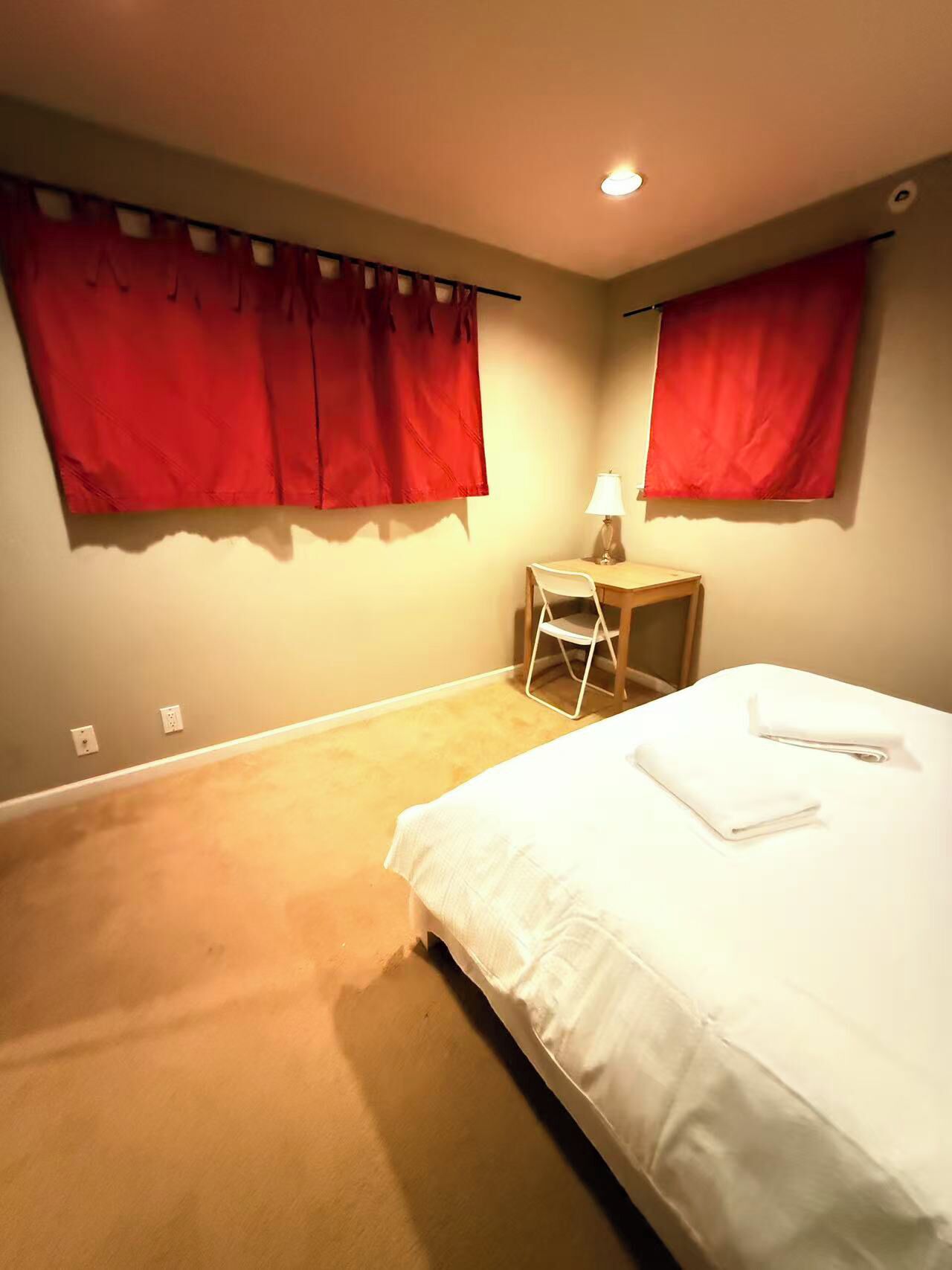 A bedroom with a bed, a chair, a small table, and two windows with red curtains.