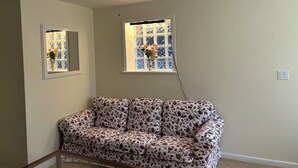 Ein Zimmer mit einer sofaartigen Sitzgelegenheit mit Blumenmuster, zwei Fenstern mit verzierten Rahmen und einer Decke mit Einbuchtungslampen.