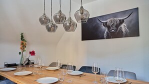 Ruokailutilassa on puinen pöytä, johon on katettu kuudelle, kirkkaat lasista valmistetut riippuvalaisimet ja seinällä on härkäaiheinen taulu.