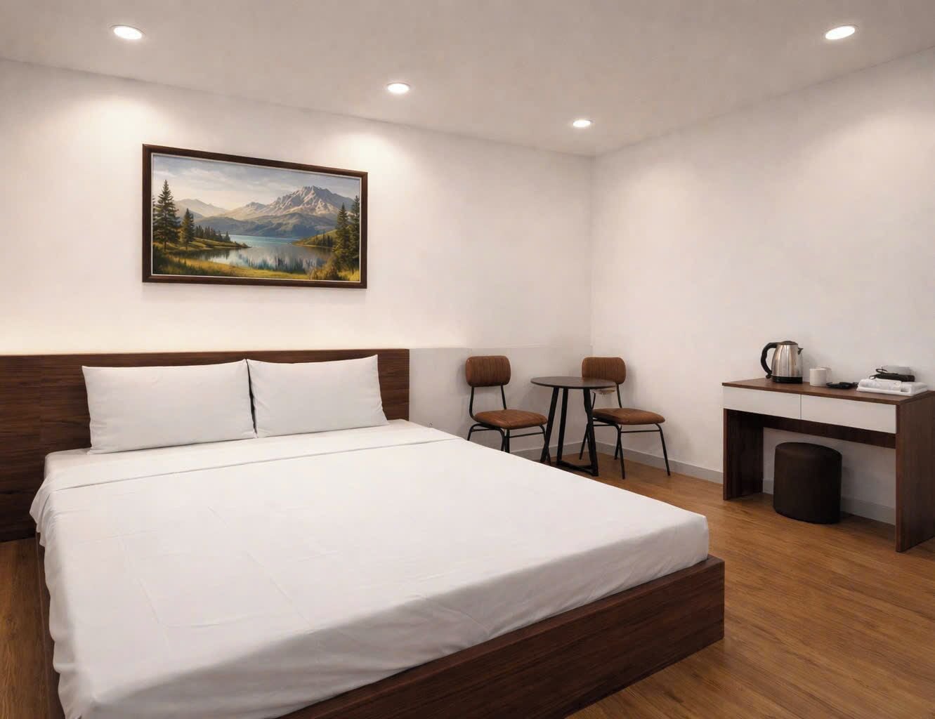 A bedroom with a bed, a desk, two chairs, a painting, and a coffee maker.