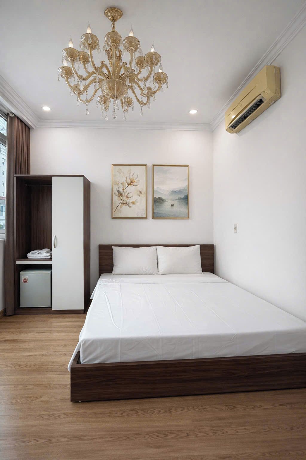 A bedroom with a bed, a wardrobe, a chandelier, and two framed pictures on the wall.