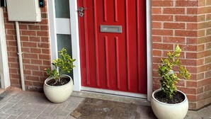 A red door with a mailbox and a number plate, flanked by two potted plants. - Perfect Spacious & Comfortable 2-bedroom Apartment in Derby for a relaxing stay (Derby)