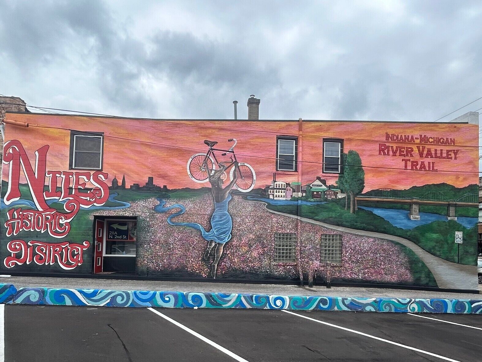 Un mural en un edificio con una sirena levantando una bicicleta, un letrero del sendero Indiana-Michigan River Valley, y un estacionamiento.
