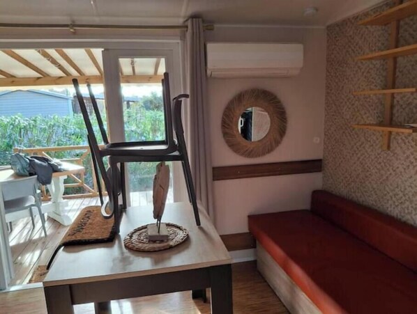 A room with a table, a chair, a red sofa, a mirror, and a window with a view of greenery. - Mobilhome in a 3* campsite in argeles sur mer sleeps 6 (Argelès-sur-Mer)
