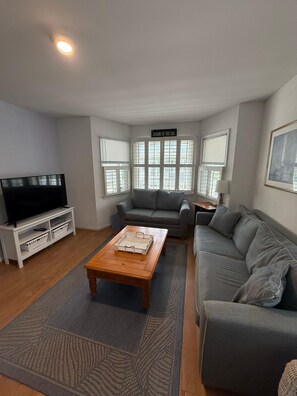 A living room with a flat-screen TV, grey sofas, a coffee table, and a sign above the windows reading 'Down by the Sea.' - Bethany Victorian Short Walk to the Beach with Wooded Yard (Bethany Beach)