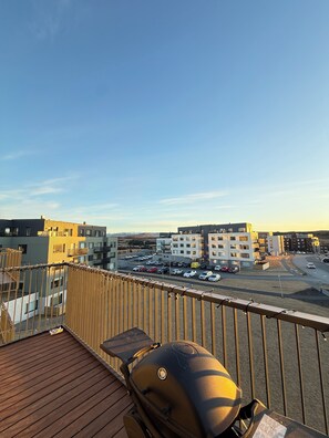 En balkong med en grill, utsikt över en parkeringsplats och moderna byggnader i bakgrunden.
