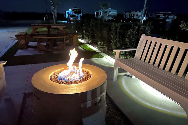 A fire pit with flames, a wooden bench, and a picnic table in an outdoor setting.
