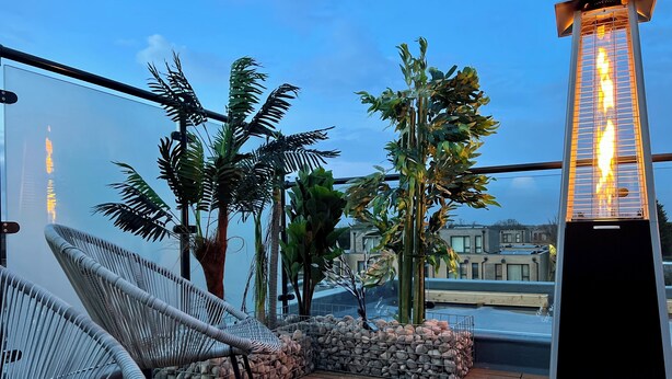 A rooftop terrace with wicker chairs, a stone planter, and a vertical fire feature.