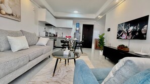 Ein modernes Wohnzimmer mit grauem Sofa, blauem Sessel, Glastisch, schwarzem TV-Schrank und einer Küche im Hintergrund.