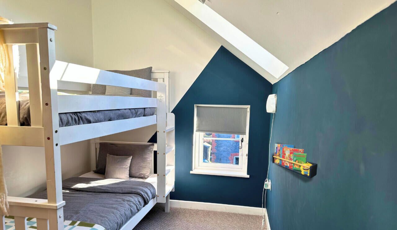 A room with a bunk bed, a window, and a wall-mounted shelf with books.