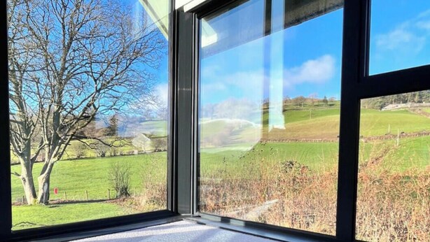 Ein Zimmer mit großen Fenstern und Blick auf eine ländliche Landschaft, darunter ein Baum und Felder.