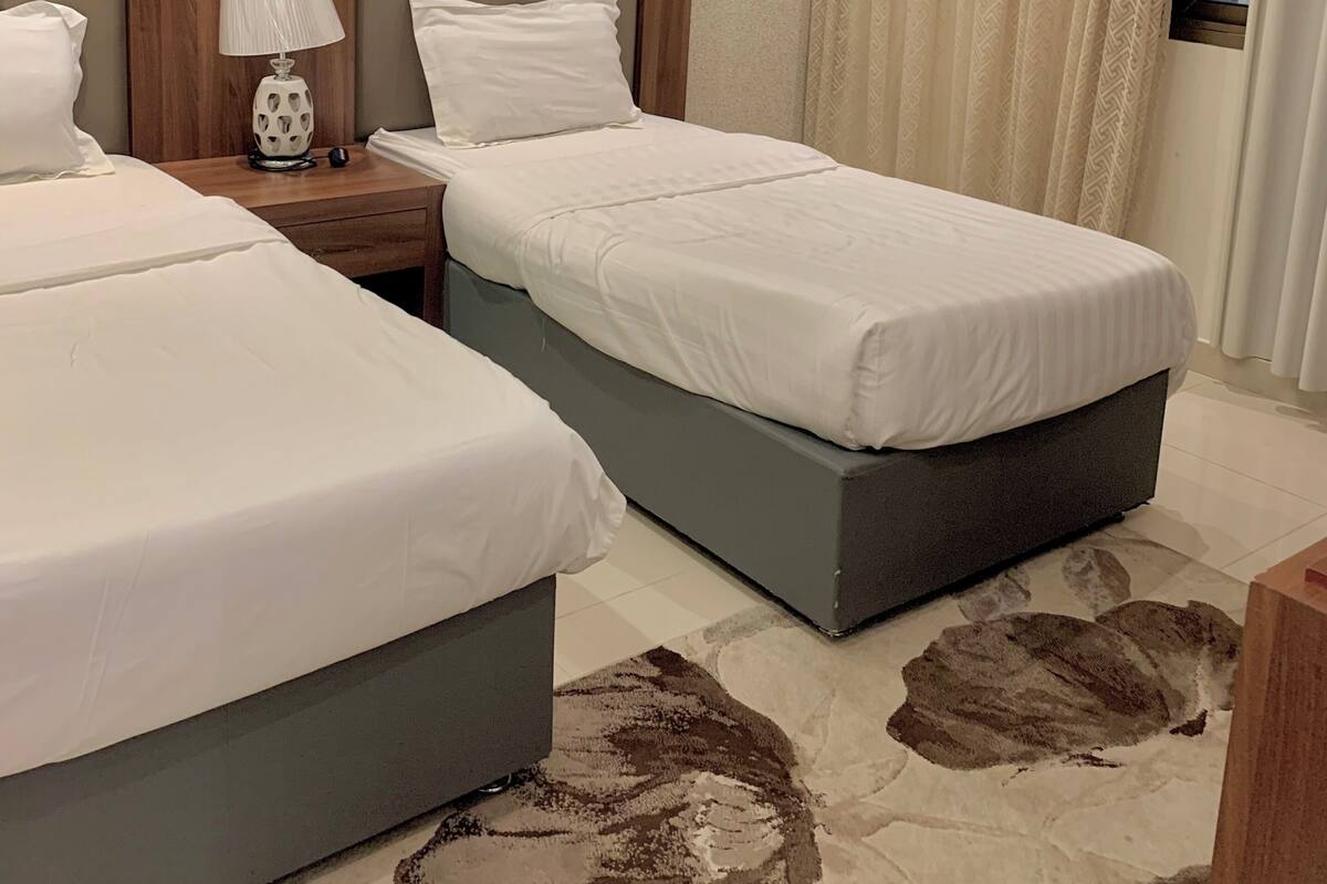 Habitación de hotel con dos camas, una mesita de noche con lámpara, aire acondicionado de pared y una ventana con cortinas.