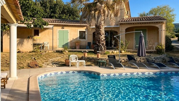 A poolside area with a palm tree, lounge chairs, and a building with a tiled roof. - Villa Du Grand Noyer (Lorgues)