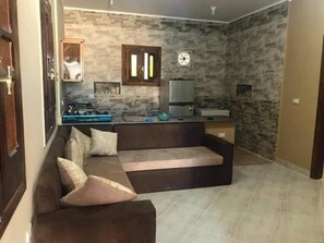 A living room with a sofa, a kitchen area with a refrigerator, and a stone wall.