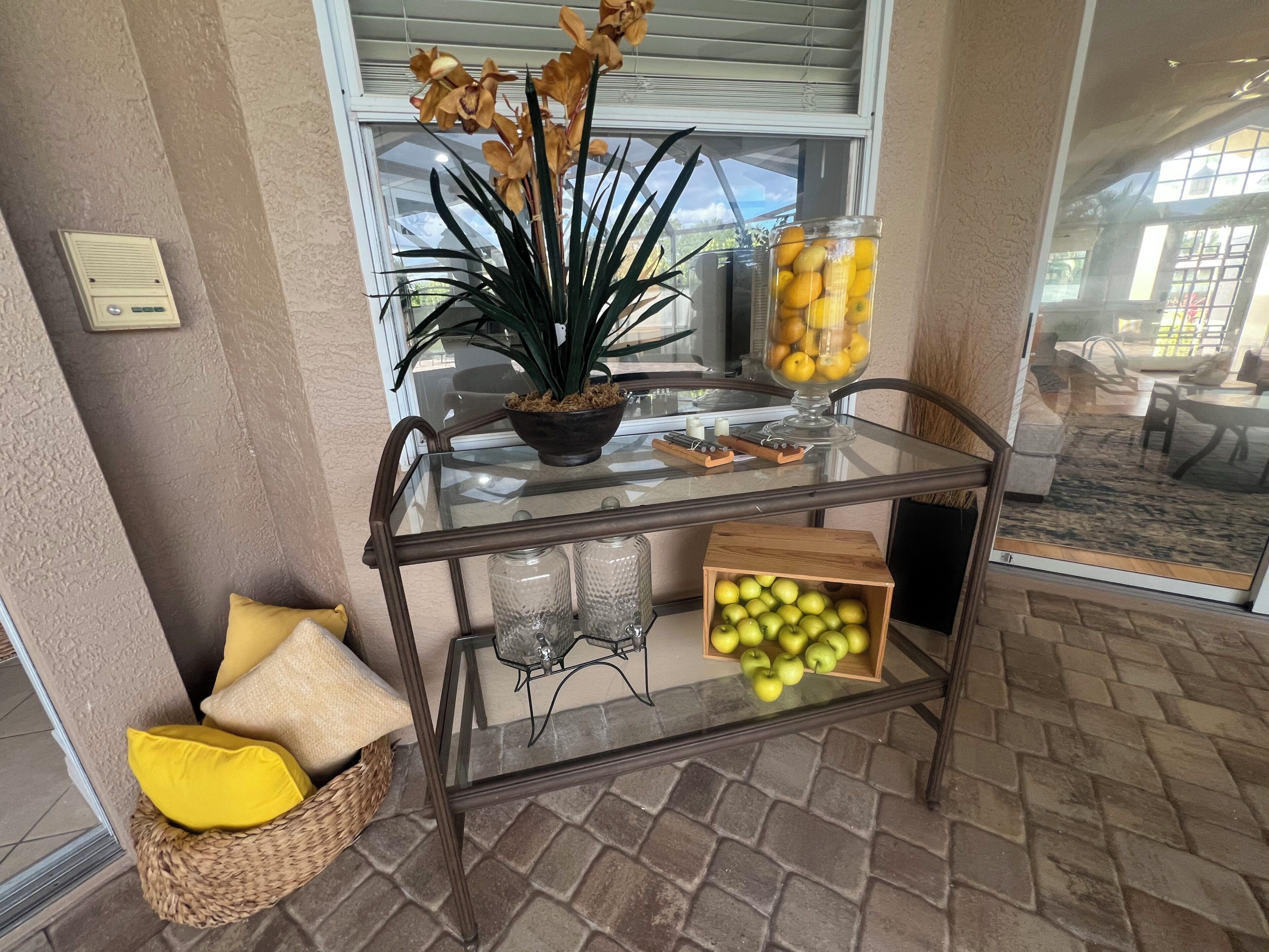 Une table dont le plateau est en verre, ornée d’une plante en pot, d’un bol de citrons et d’une boîte de pommes.