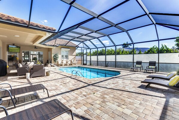 A covered outdoor swimming pool with lounge chairs, a barbecue area, and a patio with a TV.