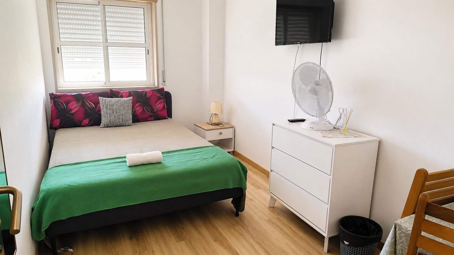 Une chambre à coucher comprenant un lit, une table de chevet, une lampe, un ventilateur et un téléviseur fixé au mur.
