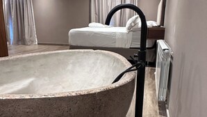 Un baño moderno con una amplia bañera de piedra, piso de madera y una cama al fondo.