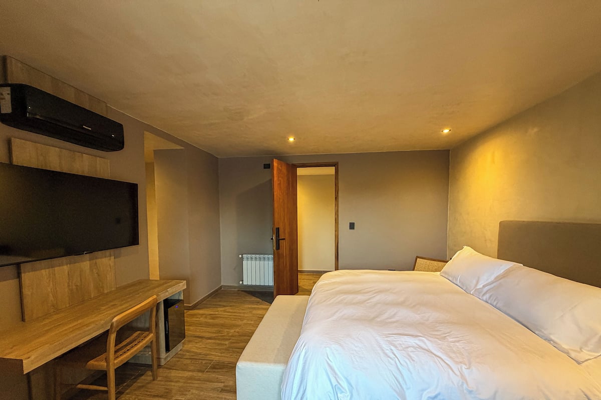 Un dormitorio con cama, televisor montado en la pared, una banca y una puerta que da a otra habitación.