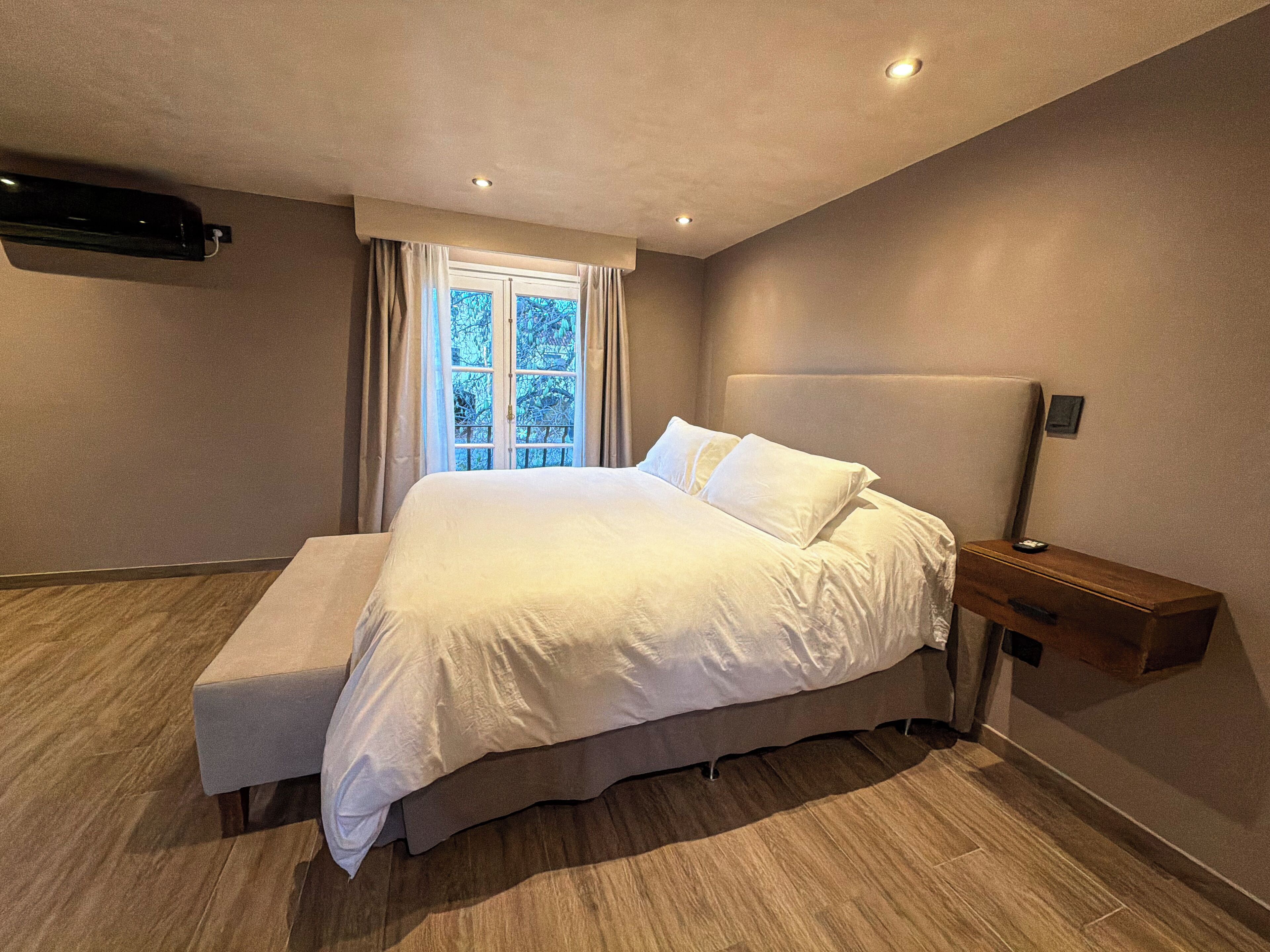 A modern bedroom with a large bed, a bench, a nightstand, and a window with curtains.