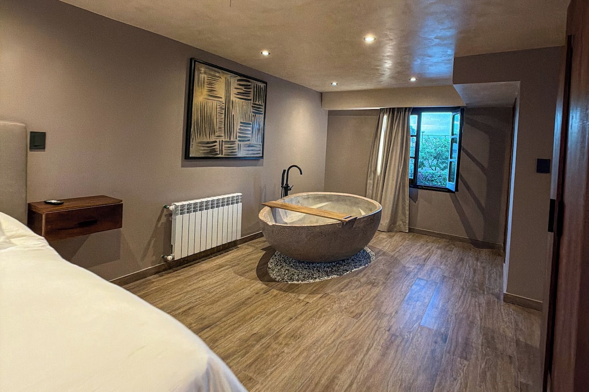 Un dormitorio moderno con una bañera grande, suelo de madera, un radiador y vistas a la vegetación a través de una ventana.