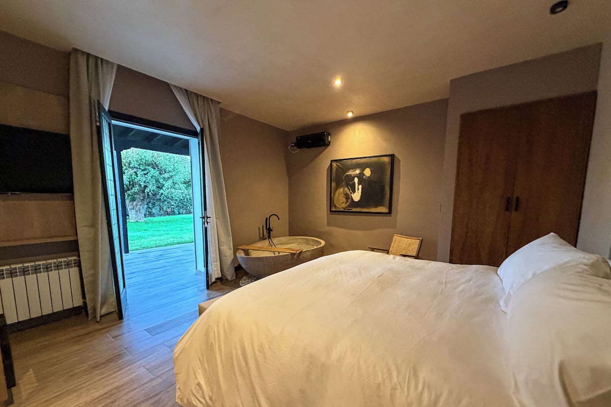 Un dormitorio con una cama grande, suelo de madera, baño con bañera y vistas a un paisaje verde a través de una puerta abierta.