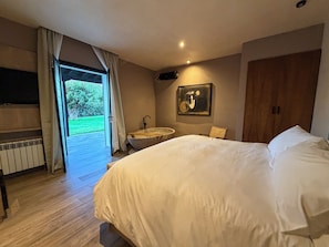 Un dormitorio con una cama grande, suelo de madera, baño con bañera y vistas a un paisaje verde a través de una puerta abierta.