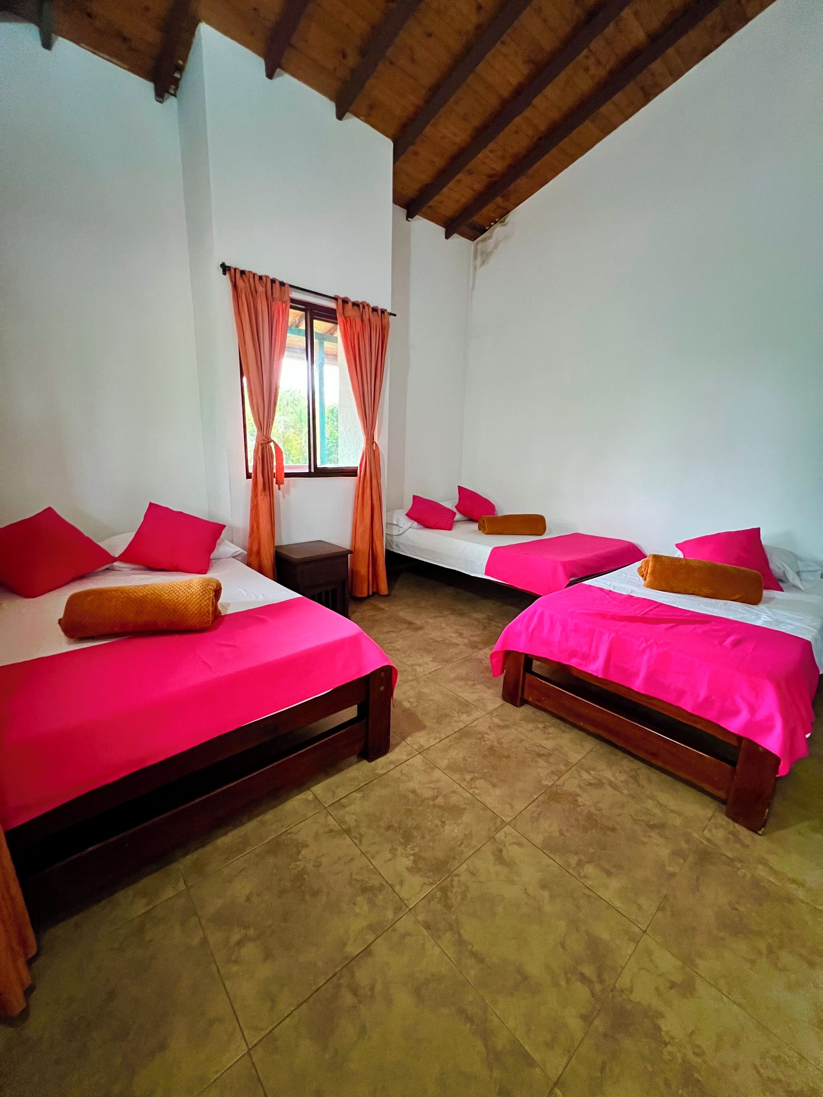 Una habitación con dos camas, cada una con estructura de madera y ropa de cama rosa. Hay una ventana con cortinas, y el suelo está cubierto de baldosas.