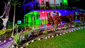 Una casa a due piani con un balcone, circondata da un giardino ricco di piante e un vialetto.