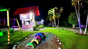 Un’area esterna illuminata di notte con un’area giochi, un edificio con tetto rosso e palme.