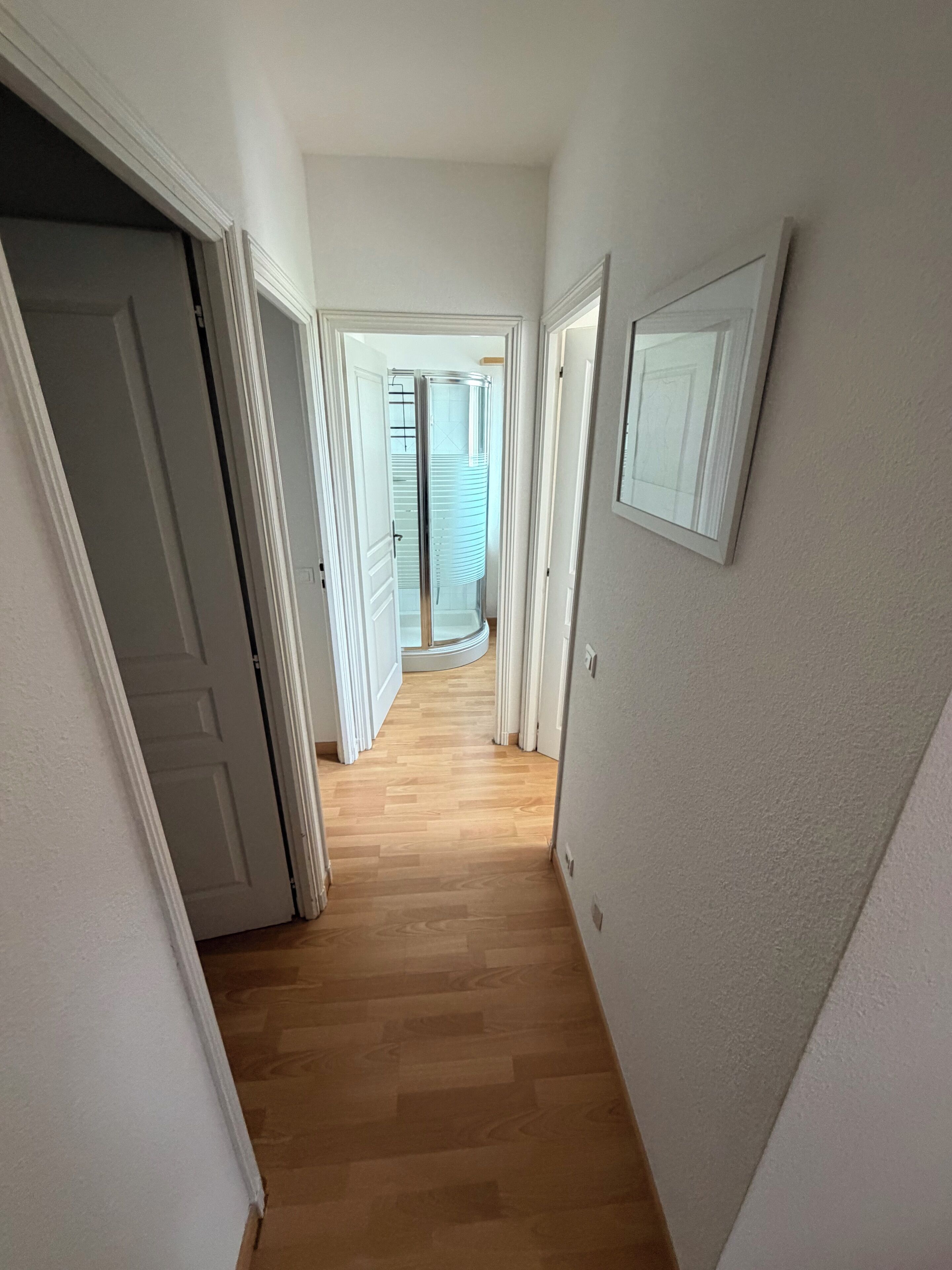 A hallway with wooden flooring, white walls, and a door leading to a room with a sliding glass door.