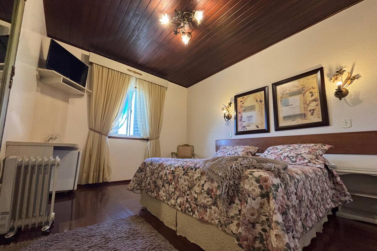 A bedroom with a wooden ceiling, a bed with floral bedding, a radiator, and two wall-mounted light fixtures.
