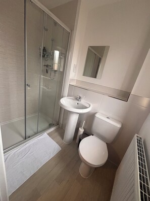 Ein Badezimmer mit Duschkabine aus Glas, einem Waschbecken auf Säule, einer Toilette und einem Spiegel über dem Waschbecken.