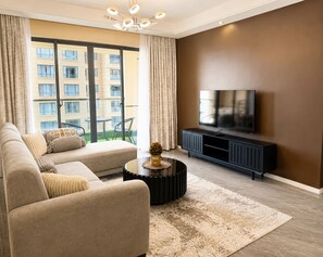 Ein modernes Wohnzimmer mit einer Couch, einem Couchtisch, einem Fernseher auf einem schwarzen Standfuß und Blick auf ein Gebäude durch große Fenster.