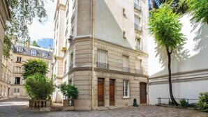A cobblestone courtyard with a building featuring a corner entrance, a small tree, and a potted plant. - A REFINED & COMFORTABLE BASE IN D MOST CAPTIVATING NEIGHBOURHOODS-SAINT GERMAIN (Paris)