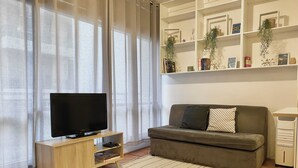 A living room with a TV on a wooden stand, a sofa with cushions, a white shelf with decorative items, and a striped rug. - The Bondi Beach Escape (Sydney)
