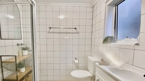 A bathroom with white tiles, a toilet, a sink, a window, and a shelf with a plant. - The Bondi Beach Escape (Sydney)