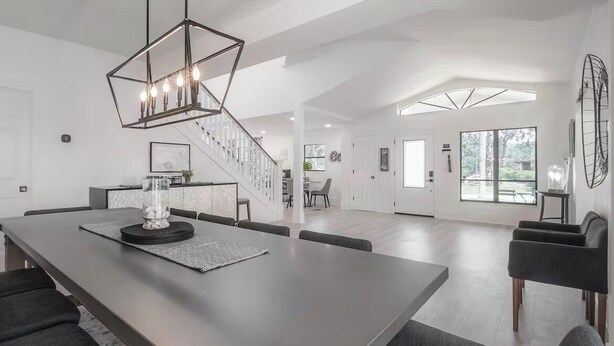 Ampla sala de estar com uma mesa de jantar central, escada e uma grande janela que ilumina o espaço com luz natural.