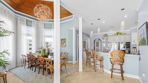 A spacious dining area with wooden chairs and a large chandelier, a kitchen with modern appliances, and a bar area with stools. - Island Girl vacation Home (Fort Myers Beach)
