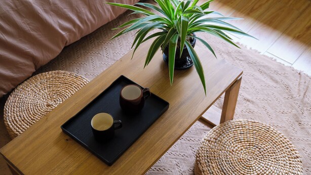 Una mesa de madera con una bandeja que sostiene dos tazas, una planta en maceta y un pouf tejido.