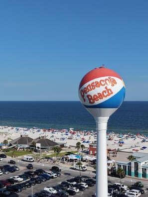 Ein Wasserturm mit der Aufschrift 'Pensacola Beach', mit Blick auf einen Strand mit Sonnenschirmen und einen Parkplatz.