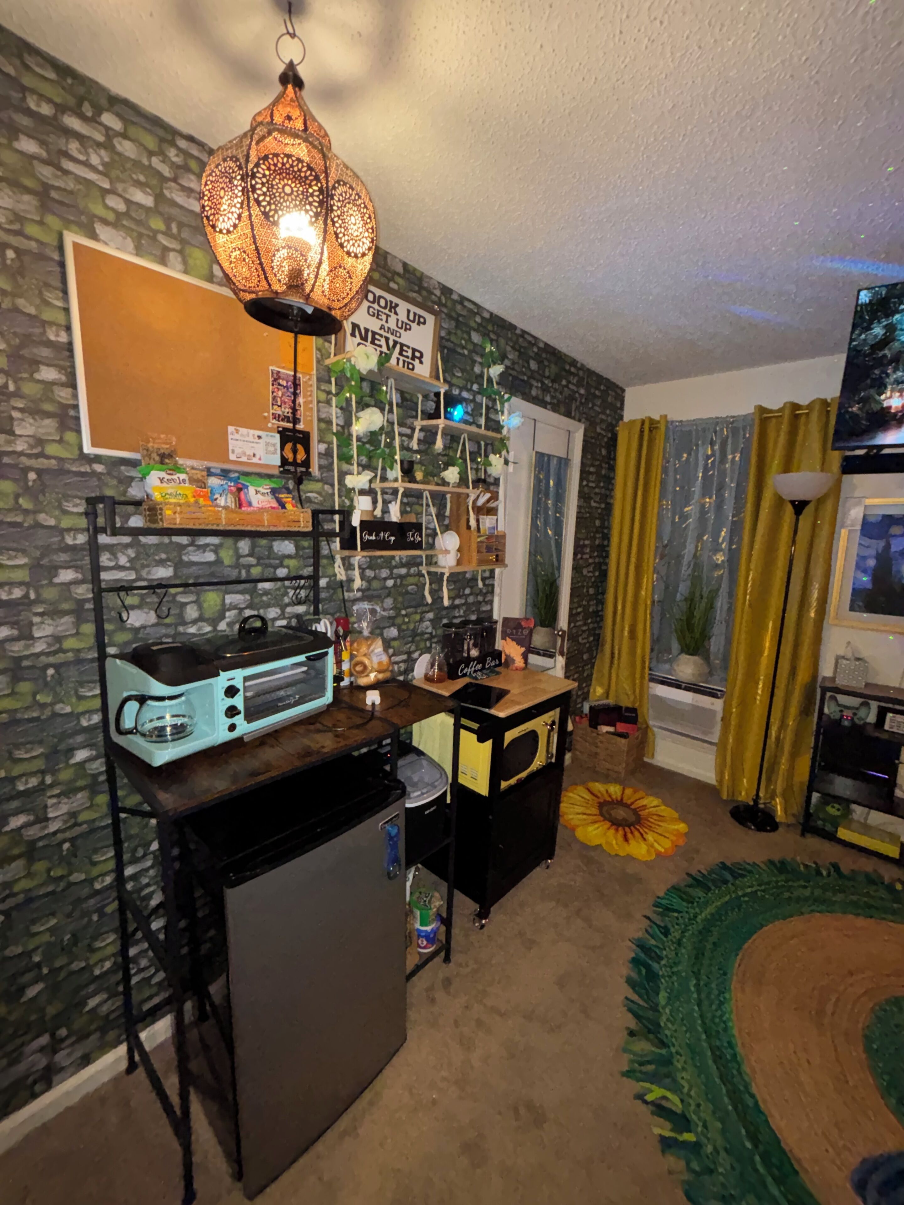 A cozy room with a coffee station, a wall-mounted shelf with various items, a hanging light fixture, and a window with curtains.
