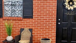 Un bâtiment en briques avec une porte noire, une fenêtre avec des volets noirs, une chaise en osier, une petite table et une plante en pot.