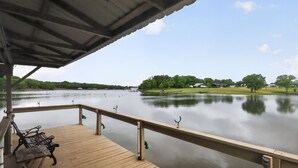 A wooden dock with a bench, a railing, and a view of a calm lake with trees and buildings in the distance. - Lake Front - Privately Owned Dock with Boat Launch (Quitman)