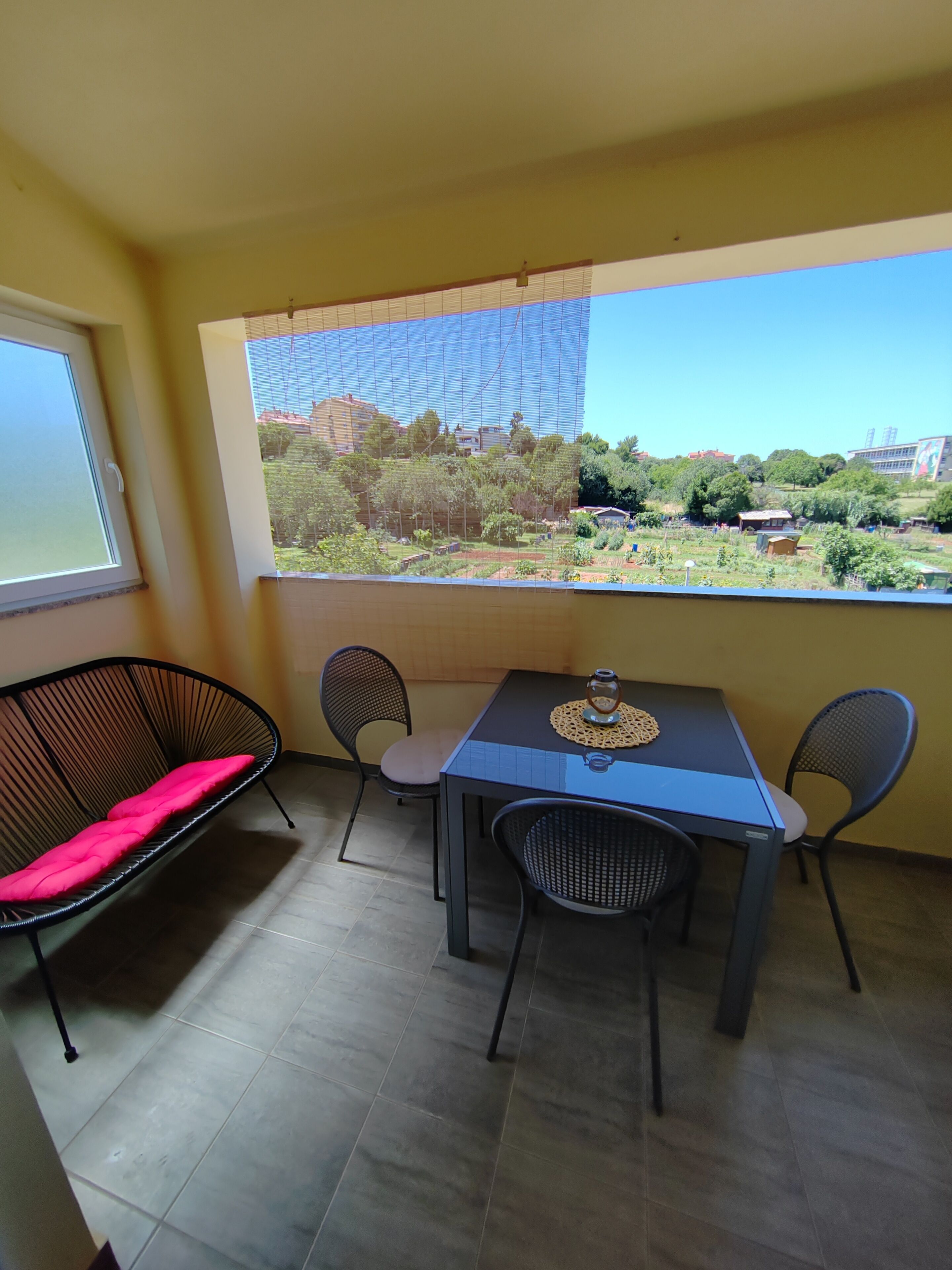 A balcony with a table and chairs, a sofa with a pink cushion, and a view of a green landscape with buildings.
