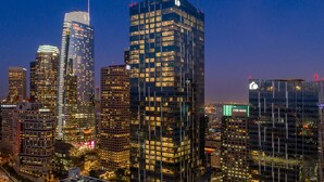 A cityscape at dusk with illuminated skyscrapers, including a prominent building with a clock tower and another with a green sign. - Glass Loft Skyline Views + Terrace (Los Angeles)