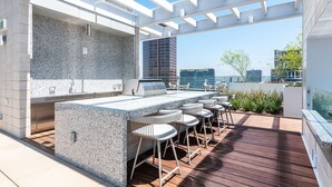 A modern outdoor kitchen with a granite countertop, bar stools, and a pergola structure. - Glass Loft Skyline Views + Terrace (Los Angeles)