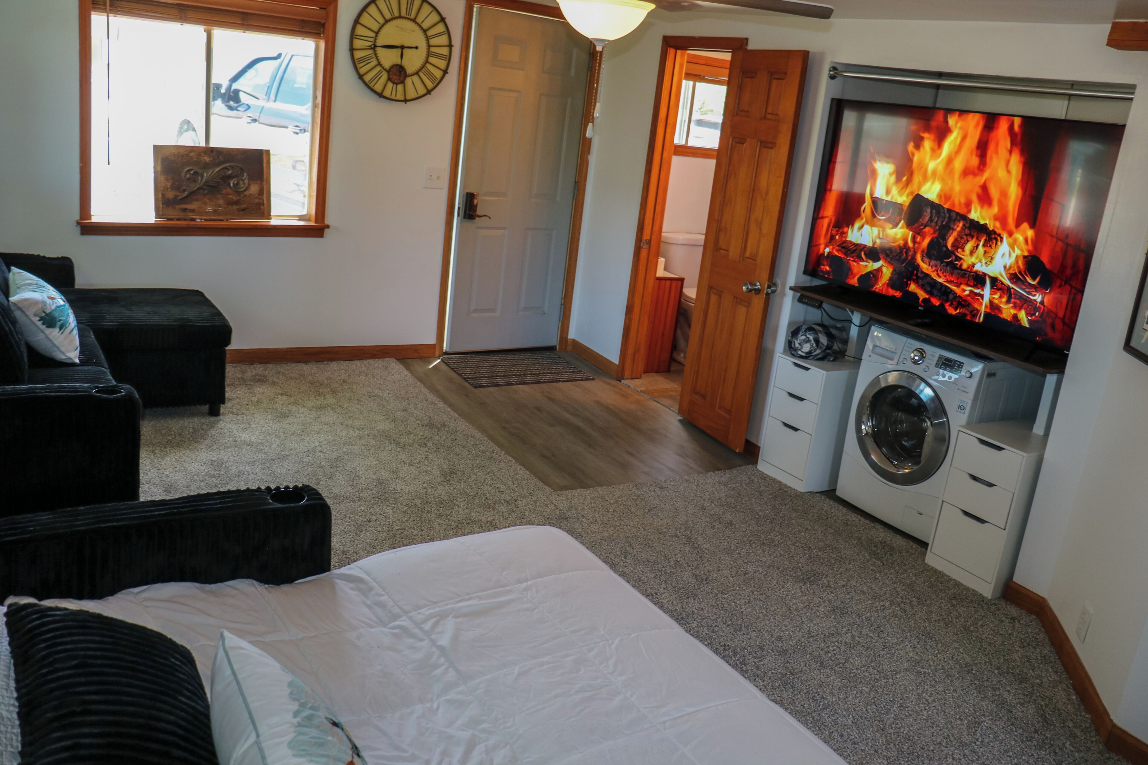 A bedroom with a bed, a washing machine, a TV displaying a fire, a clock, and a door leading to another room.