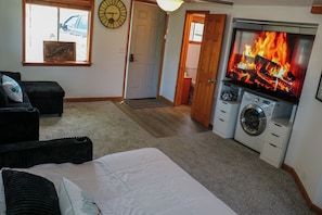 Une chambre à coucher avec un lit, un lave-linge, un téléviseur diffusant une image de feu de cheminée, une horloge et une porte donnant sur une autre pièce.
