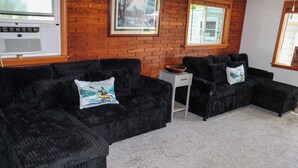 A living room with a brick wall, two black sofas with cushions, a small white side table, and a window with blinds.
